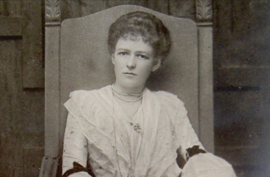 Black and white photo of a young woman sitting in a chair. She is wearing a lace dress and has a thoughtful expression.