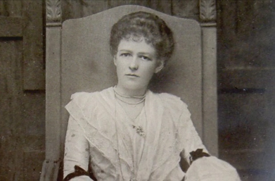 Photo en noir et blanc d’une jeune femme assise sur une chaise. Elle porte une robe de dentelle et semble songeuse.