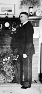Black and white photo of a man wearing a suit, standing in front of a fireplace.