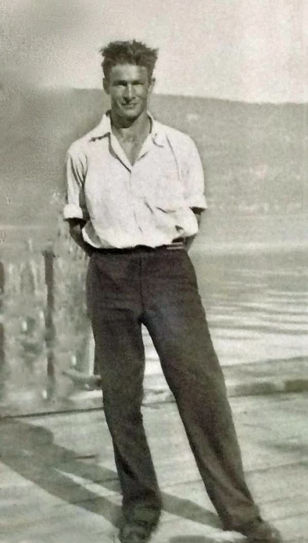 Black and white photo of a young man standing on a dock, lake in the background.