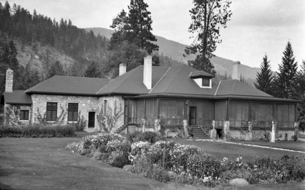 Black and white photo of a one-level large stone house with a screened porch.