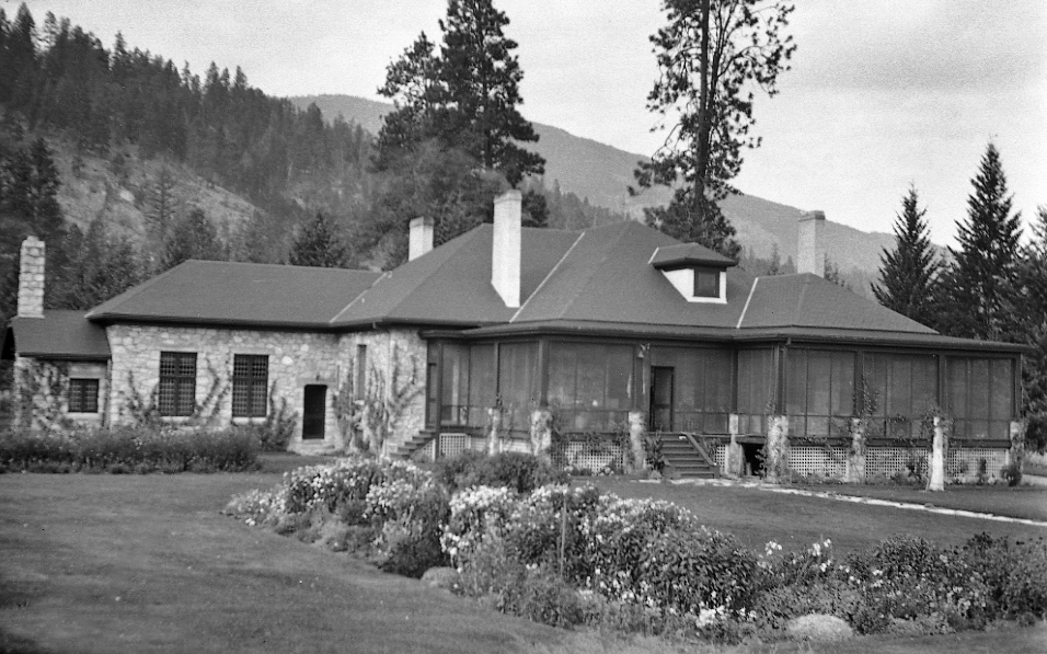 Black and white photo of a one-level large stone house with a screened porch.