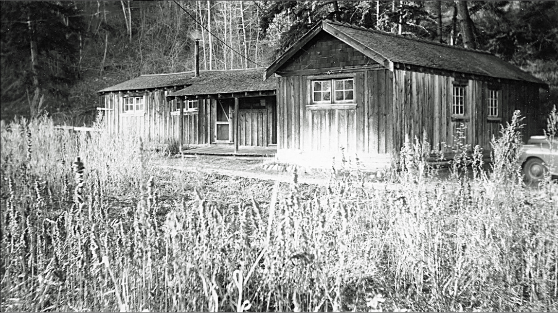 Black and white photo of a wood-sided building. Tall grasses in the foreground.