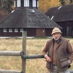 Colour photo of a man wearing a hat and smoking a pipe, leaning on a fence near a round barn.