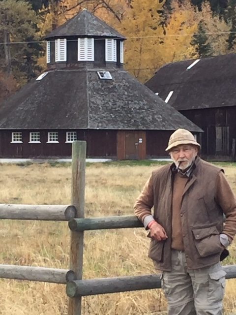 Colour photo of a man wearing a hat and smoking a pipe, leaning on a fence near a round barn.