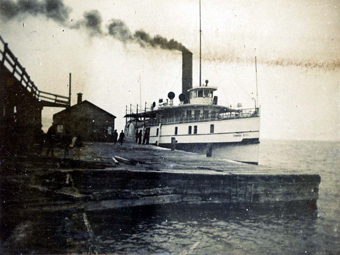 Black and white photograph of a steamboat docked at pier. Small building on the pier to the left of the boat, and a small group of people on the pier beside the boat.