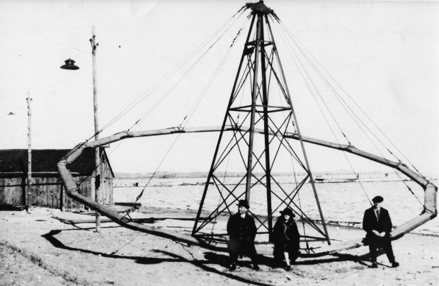 Black and white photograph of three people sitting a circular swing the lake and pier and are in the background.