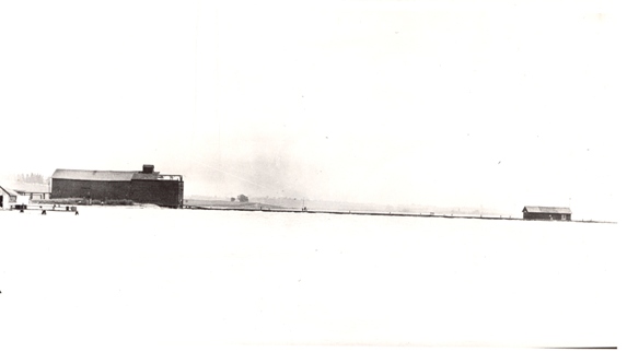Black and white photograph of a large storage shed located left of the pier leading to smaller shed.