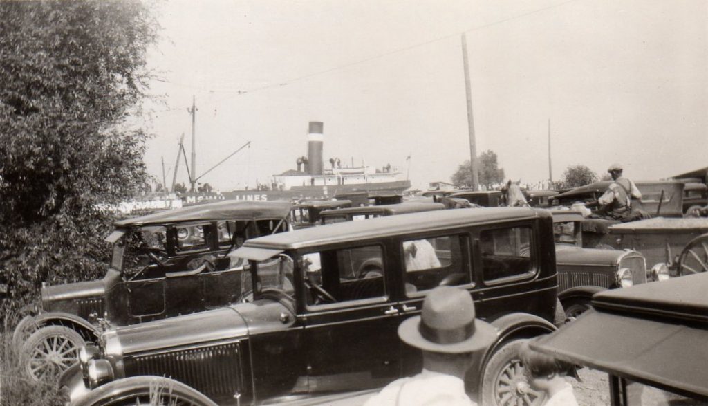Black and white photograph of old cars parked with people standing around them. A steamboat is visible in the background.
