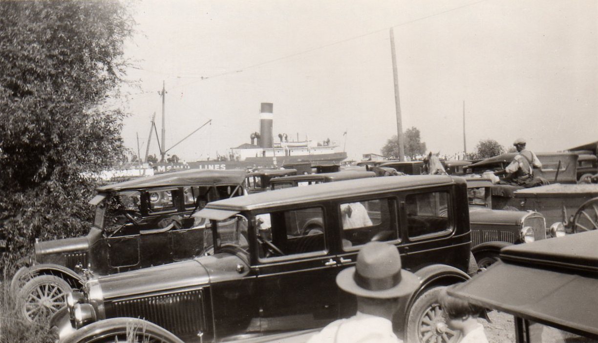 Black and white photograph of old cars parked with people standing around them. A steamboat is visible in the background.