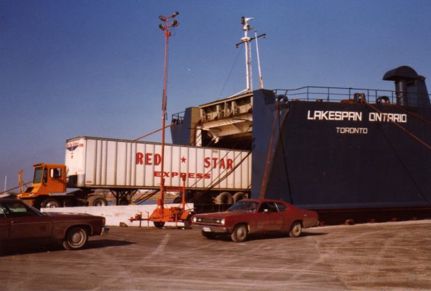 Colour photograph of red and white transport truck, backing into large ship, Lakespan Ontario, that is docked at the pier.