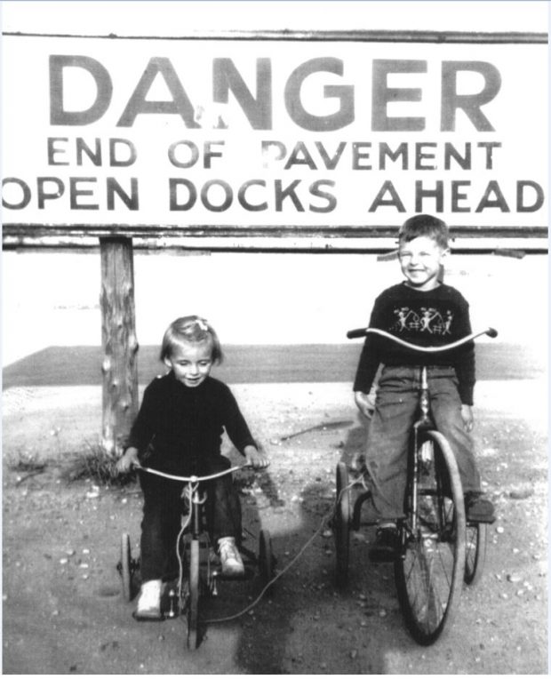 Black and white photograph of two children on tricycles by a large sign that says Danger End of Pavement Open Docks Ahead.