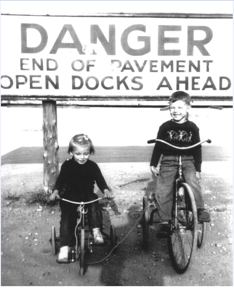 Black and white photograph of two children on tricycles by a large sign that says "Danger End of Pavement Open Docks Ahead".