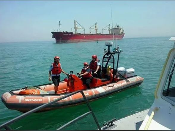 Colour photograph of three people in helmets and life vests in a small orange Coast Guard rescue boat on the water. Larger red ship in the background.