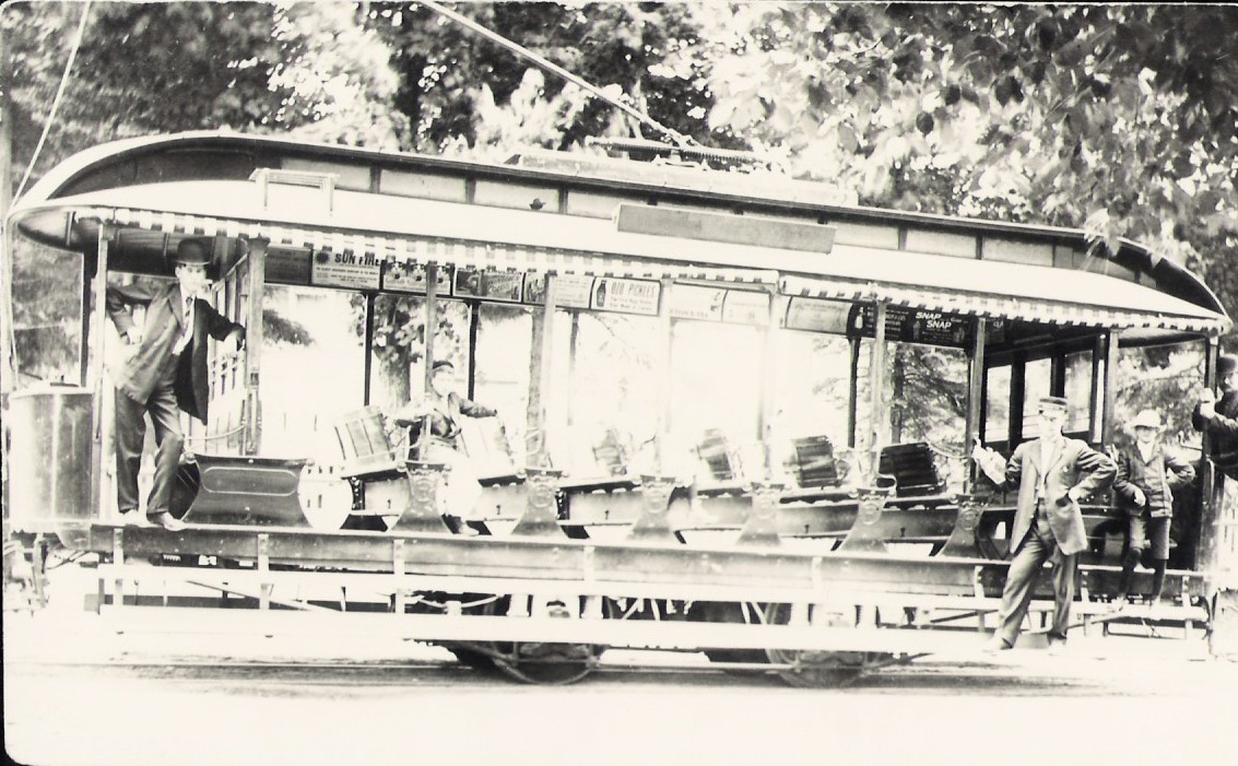 Black and white photograph of a street car with open sides and benches in middle. Three people standing and leaning on the street car and one person sitting on seat.