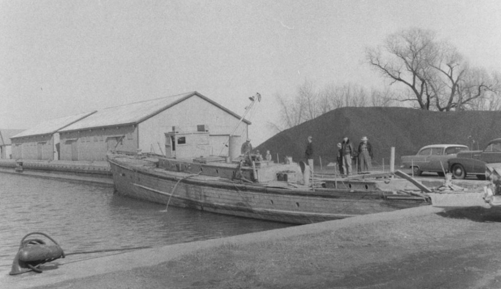 A group of individuals standing near a boat, Harry H, docked at the Oshawa Harbour.