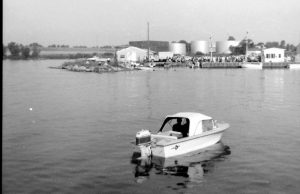 Black and white photograph of a small motor boat on the water. In the background are buildings, boats, docks, and oil tanks.
