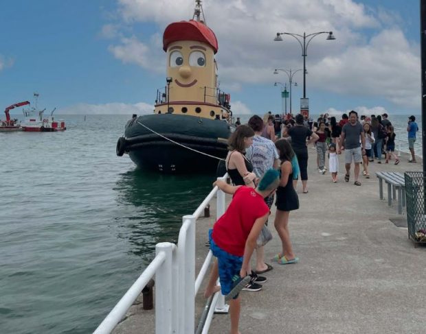 Colour photograph of a tug boat docked at a pier where a large group of people is standing.