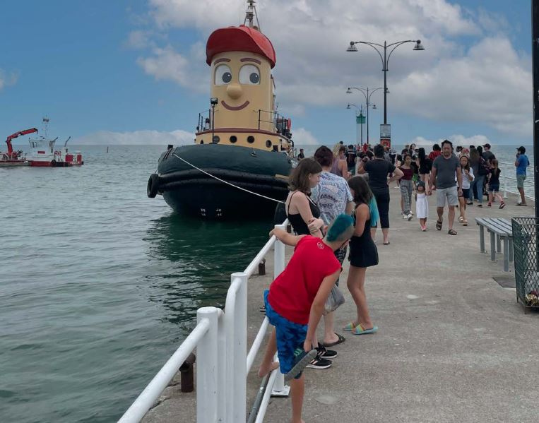 Colour photograph of a tug boat docked at a pier where a large group of people is standing.