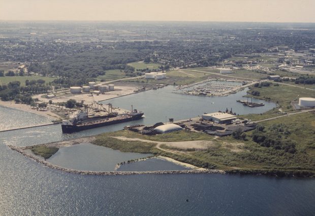 Colour aerial photograph of the Oshawa Harbour lands. There is a large ship docked in the main channel, a few harbour buildings in the centre of the image and a view of the marina in the background.