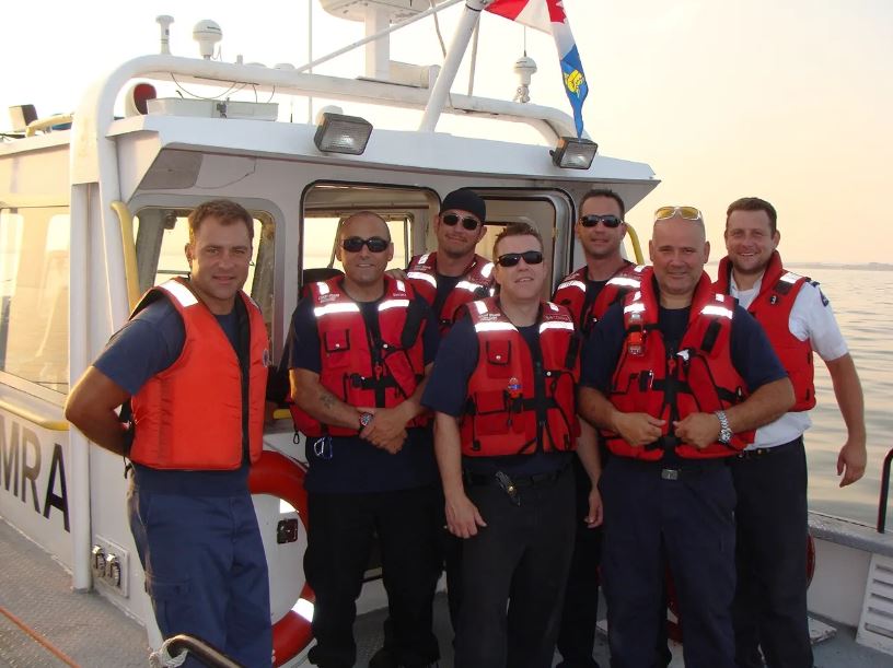 A colour photograph of a seven people wearing red life jackets standing on a boat.