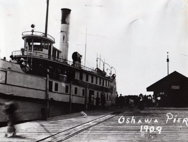 Black and white photograph of a steamboat docked at pier. Small building on the pier to the right of the boat, and large group of people standing on pier beside boat.