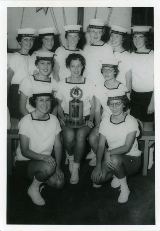 Black and white photograph of eleven people around a trophy. They are all wearing white shirts with the same caps.