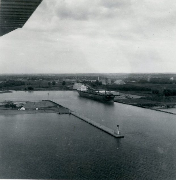 Black and white aerial photograph of a harbour with a pier and a large ship docked on the east side.