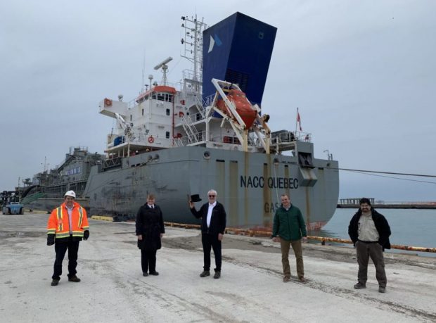 Colour photograph of five people, one is holding a top hat, standing in front of a large ship.