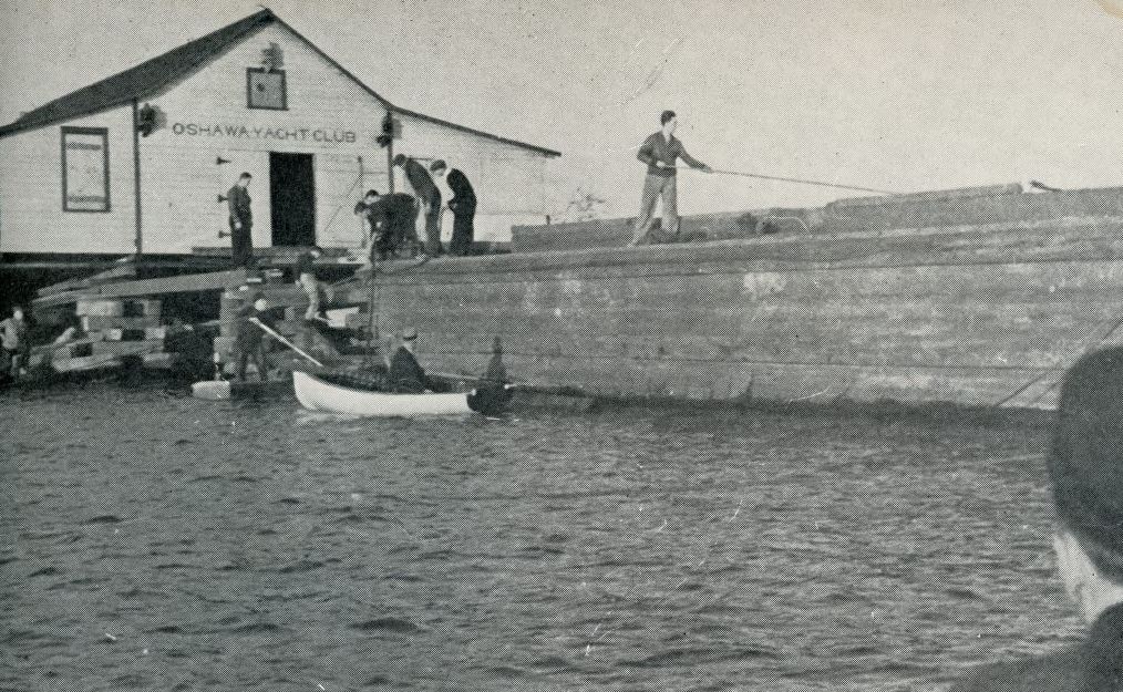 Black and white photograph of a large yacht club building. A group of people is around the building and a smaller boat is in the water.