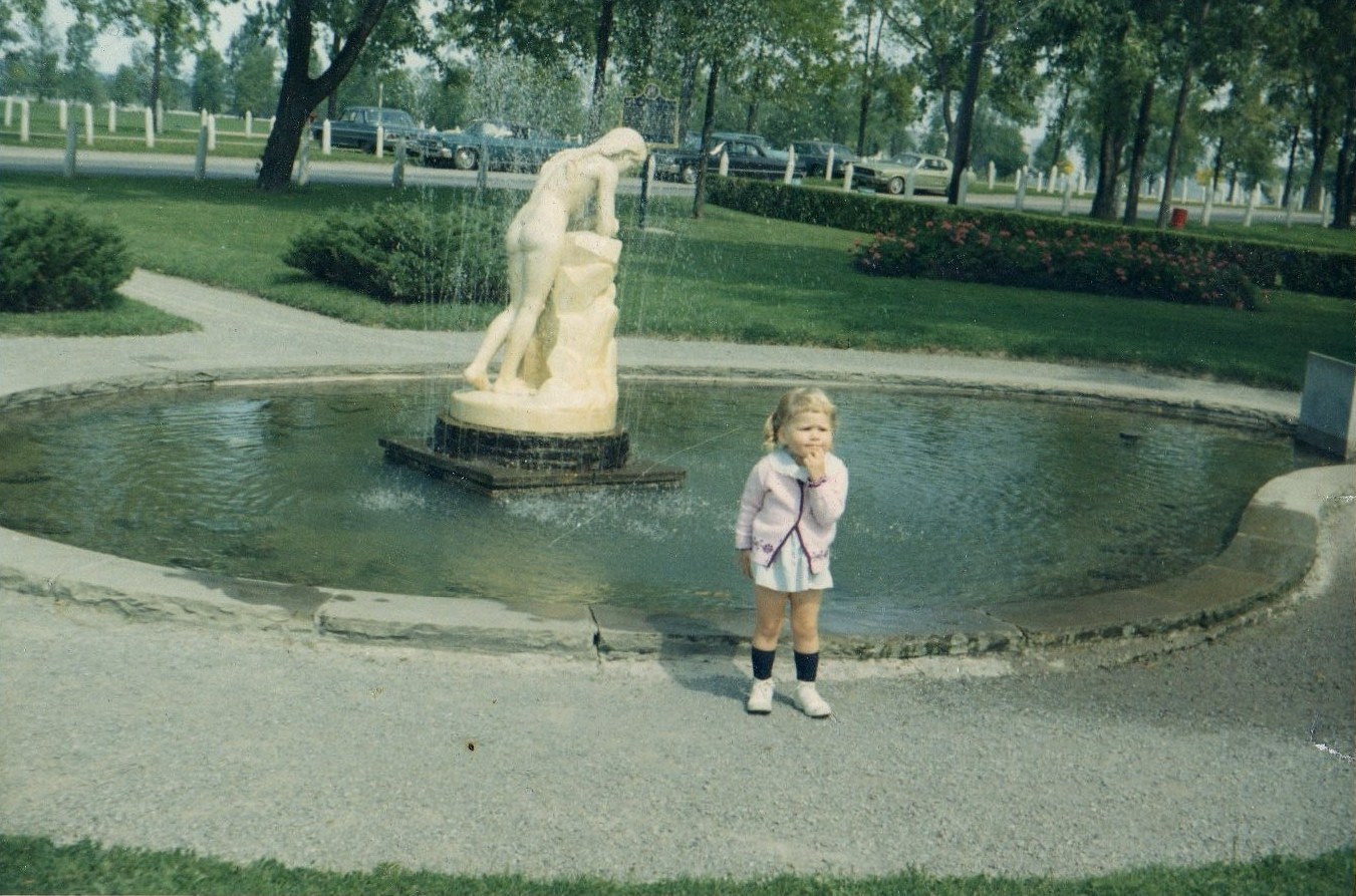Colour photograph of a child standing in front of marble statue surrounded by a pool of water. Trees line the street behind the statue and cars are parked on the street.