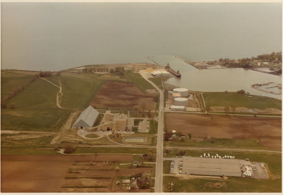 Colour aerial photograph of the land around the Oshawa Harbour. There are two large factories and a ship unloads raw sugar at the harbour.