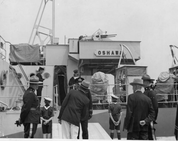 Black and white photograph of boat, H.M.C.S. Oshawa, with people in uniforms and suits on the deck of the boat and standing on the pier beside boat.