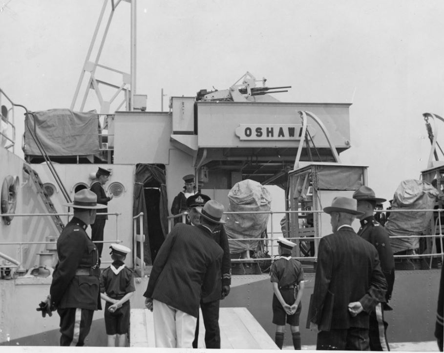 Black and white photograph of boat, H.M.C.S. Oshawa, with people in uniforms and suits on the deck of the boat and standing on the pier beside boat.
