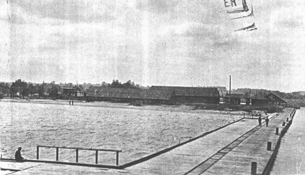 A copy of a black and white photograph of a wooden pier, and people are on the pier walking. Other buildings are in the background.