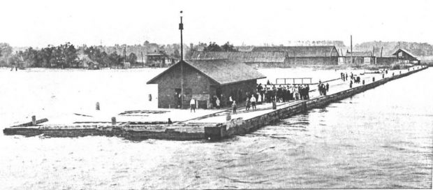 Black and white copy of the people and a small building standing on a wooden pier. Other buildings are in the background.