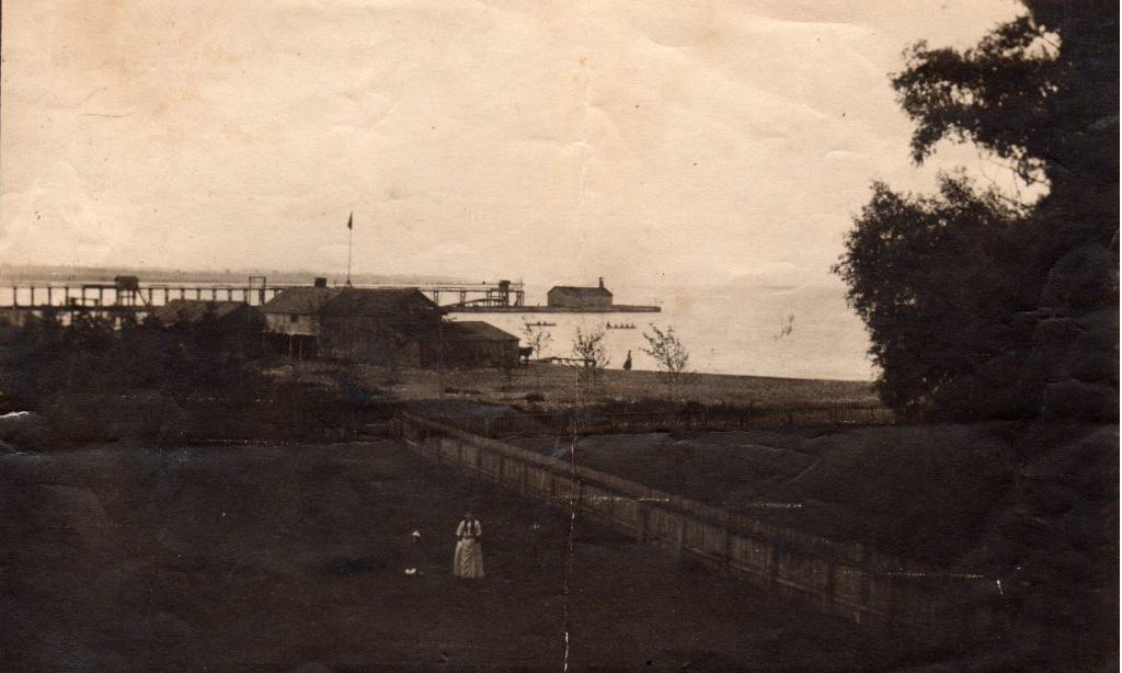 Black and white photograph of a person in a field with large buildings in the distance. The pier and lake are visible in the background, behind the buildings.