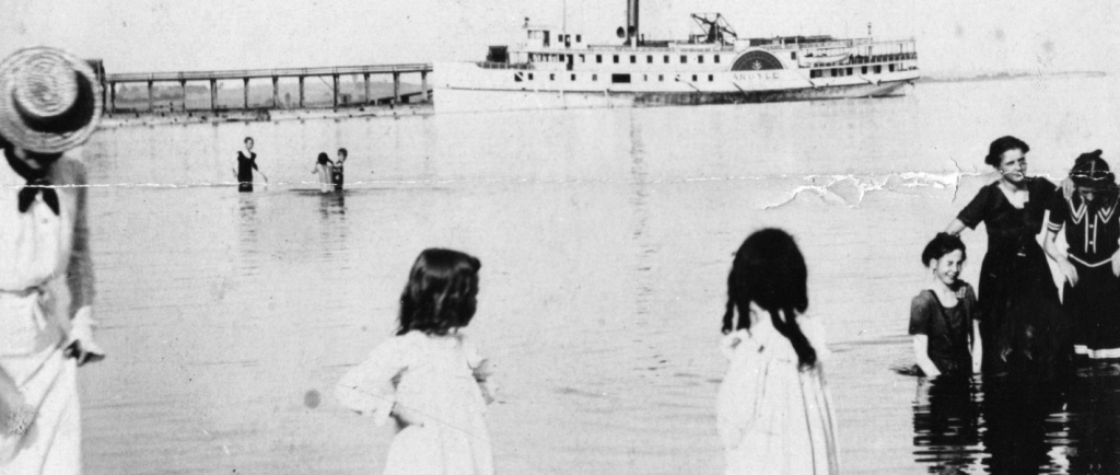 Black and white photograph of a lake with individuals standing in the water and in the background a large steamship and peir.