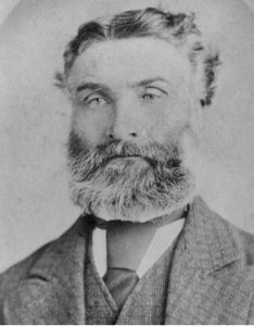 A black and white photograph of a person with a beard wearing a suit jacket with a tie.