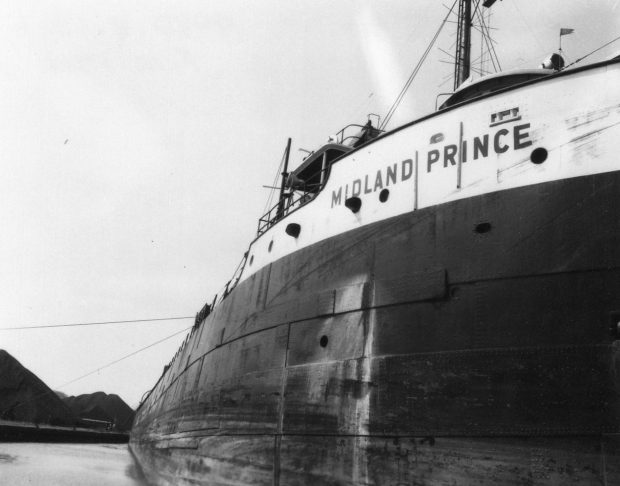 Black and white photograph of a large ship. Midland Prince is written on the side of the ship. Coal piles are in the background.