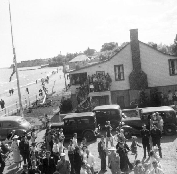 Black and white photograph of a group of people at the lakefront. Cars and a building with a large anchor out front.