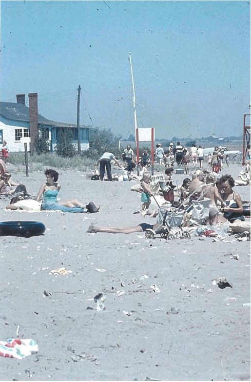 Colour photograph of several people sitting on the beach with a house in the background.