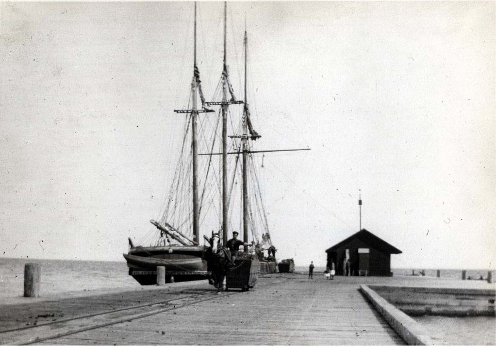 Black and white photograph of a schooner docked at pier with a small building on the right of the pier. There is a person with a horse and cart beside the schooner on the pier.