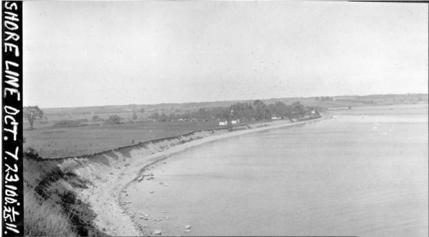 Black and white glass slide of a shoreline with a large body of water. On land there are buildings and trees.