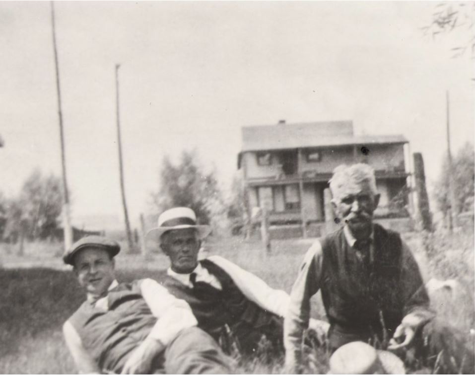 Black and white photograph of the three people sitting in the grass. A large two storey house is in the background.