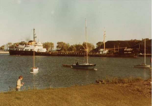 Colour photograph of a large ship docked at the harbour unloading coal. A person is seen near the bottom of the image and small boats are anchored in the water.