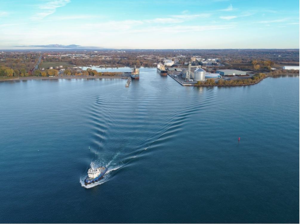 Colour aerial photograph of a harbour with two ships docked on the east and west wharf. A tug boat departs the harbour.