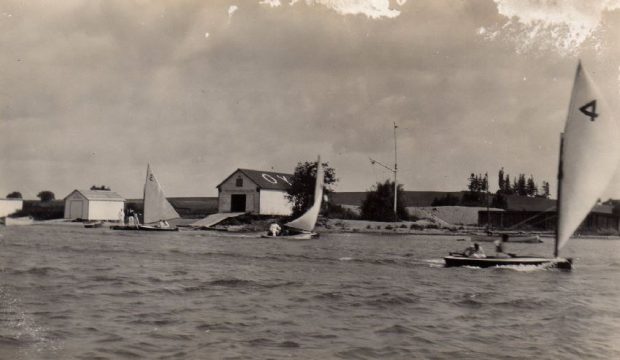 Black and white photograph of a large yacht club building with two smaller buildings on the right. All three buildings sit along the lakefront.