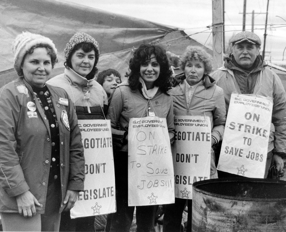 Four women and one man stand in front of a tarp in winter clothes. They wear picket signs reading “Negotiate Don’t Legislate” and “On Strike to Save Jobs”. A young boy can be seen looking out between two of the women.