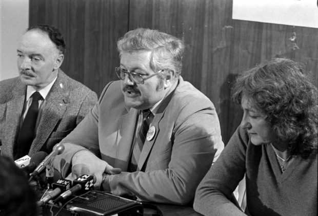 Two men and one woman sit at a table. Four microphones and a tape recorder are in front of them.